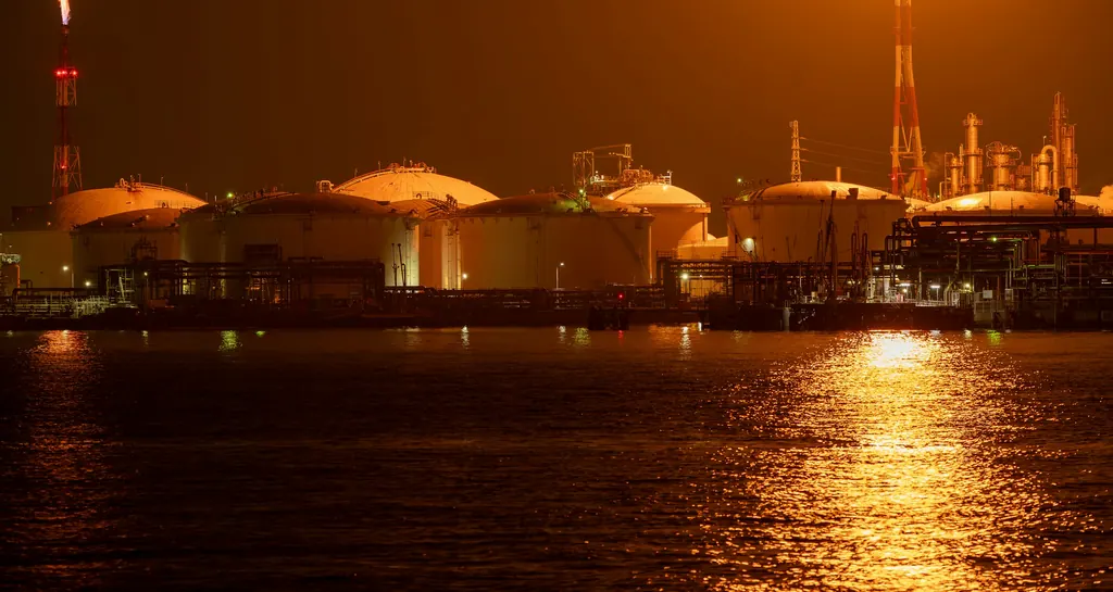 Oil storage tanks lit at night beside the water, their lights reflected across a calm harbour